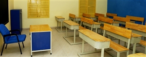 A classroom with a blue chair to the left in front of a wood desk with blue sides and long wooden desks with chairs for the students. Photo by  Haseeb Modi via Unsplash.